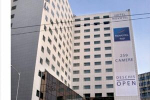 Hotel Novotel, wysoki budynek, biały blok, niebieskie logo, napis Open